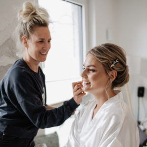 Bridal Stylistin schminkt blonde Braut. Die Braut sieht man von der Seite